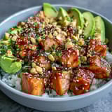 A close-up of a vibrant Avocado Salmon Bowl shows chili oil drizzle, chopped peanuts, nori strips, and a bright green dollop of wasabi paste.