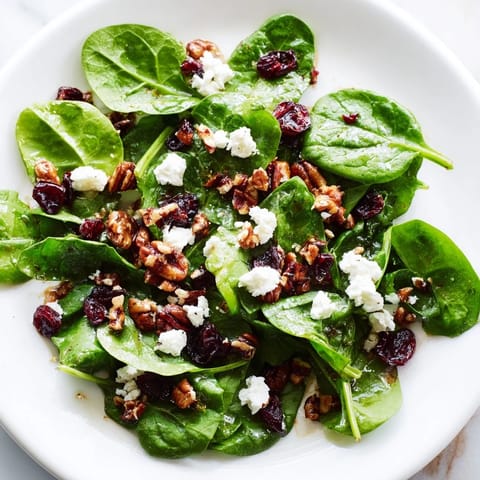 A vibrant spinach goat cheese salad with creamy crumbles, sweet dried cranberries, and candied pecans on a white platter.