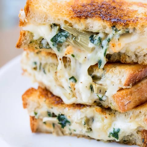 A close-up of the Artichoke Spinach Grilled Cheese reveals gooey cheese stretching between crispy, buttery bread slices.