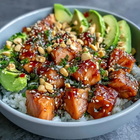 A close-up of a vibrant Avocado Salmon Bowl shows chili oil drizzle, chopped peanuts, nori strips, and a bright green dollop of wasabi paste.